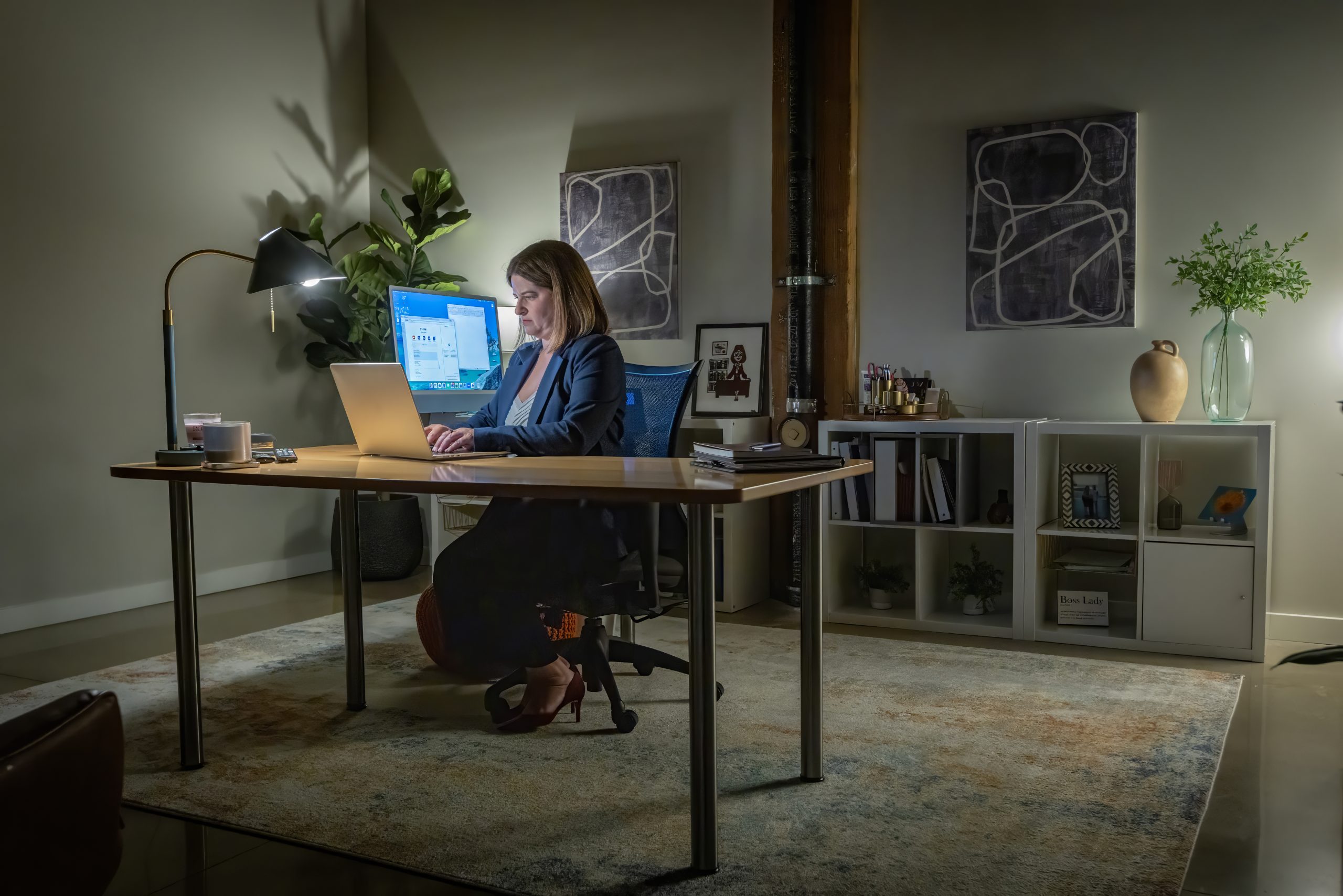 7E0A8647 - West Michigan Divorce Woman in a blazer typing on a laptop at a home office desk with a monitor, lamp, and plants nearby.